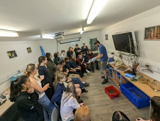 A photograph of a group of people in a classroom listening t someone talking