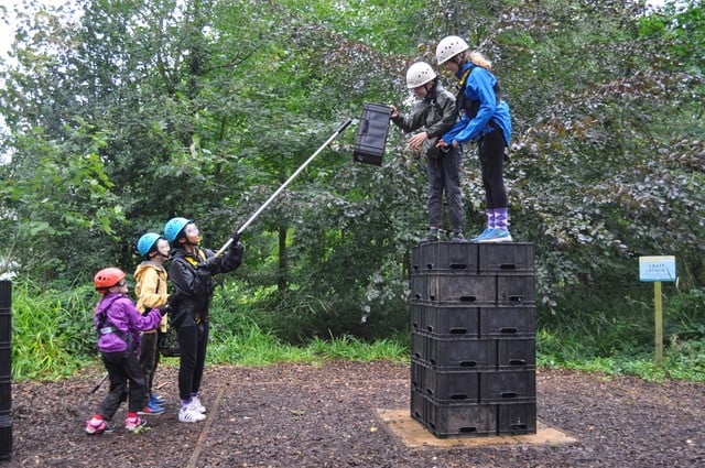 Children working together to build a stack of crates. " children stood on top while 3 pass up the crates with a long stick