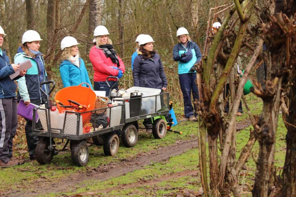 8 people dressed in outdoor clothing and hard hats watching a forest school demonstration