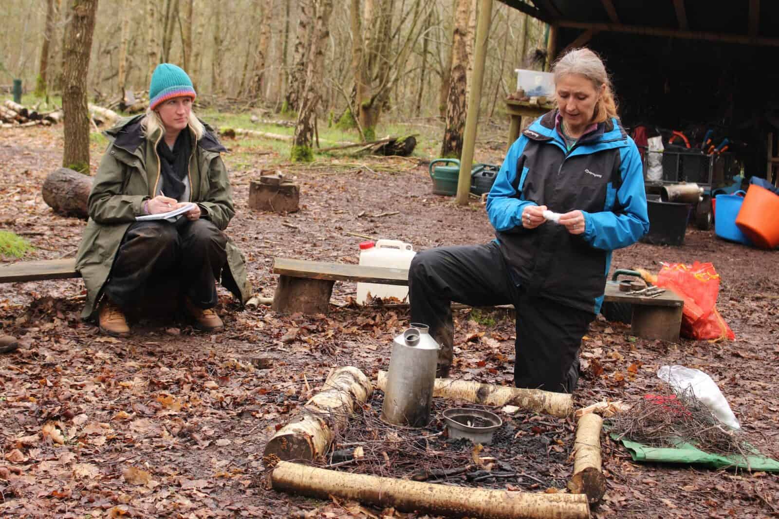 Two women in outdoor clothing, participating in a demonstation of safely usin a kelly kettle
