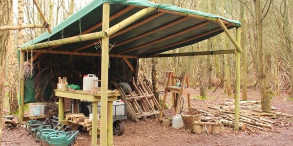 Outdoor classroom structure in a wood with a green canvas roof and contains forest school equipment