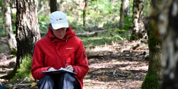 woman in red jacket in the woods taking notes