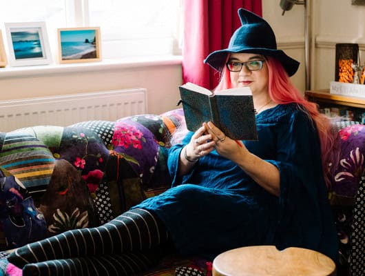 A photograph of a women sat on the sofa in a witches hat, reading a book