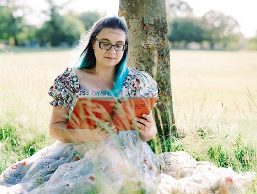 A photograph of a women sat in a field reading a book