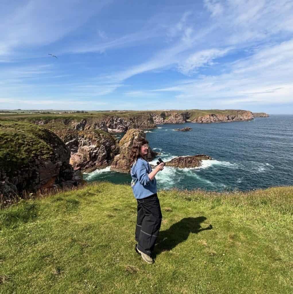 A woman in a blue sweatshirt and black trouser is on a coastal cliff holding binoculars.
