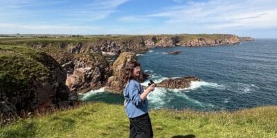 A woman in a blue sweatshirt and black trouser is on a coastal cliff holding binoculars.