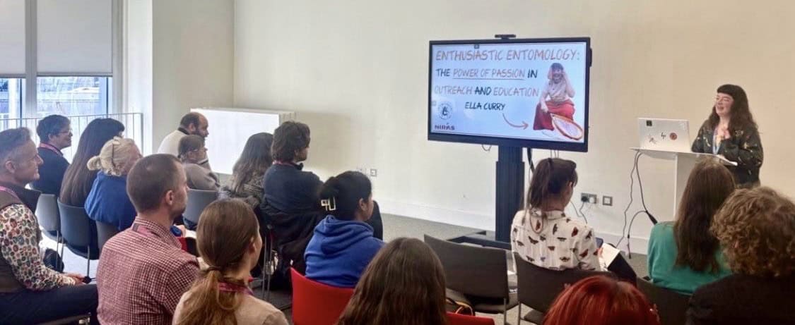 A woman delivering a presentation in front of a group of people. A slide on a screen reads ' Enthusiastic Entomology - The power of passion in outreach and education by Ella Curry'